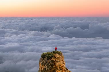 A hiker stands at the top of a mountain looking down over clouds and a sunset