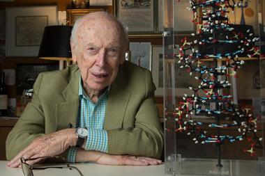 A photograph of James Watson sitting next to a molecular model of DNA's double helix