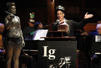 Marc Abrahams in his signature top hat presents the Ig Nobel prize ceremony. On stage with him is a woman in silver body paint wearing a chainmail bikini and holding a torch.