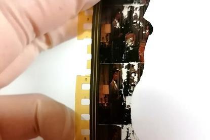 Close up of a damaged strip of celluloid film held by a hand in latex gloves. The film features two women in mid-century living room and clothing but the film is cracked and broken and parts of the images have turned white.