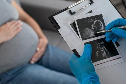 Pregnant woman in the background sitting on a couch while a doctor looks at an ultrasound image of a fetus on a clipboard in the foreground.