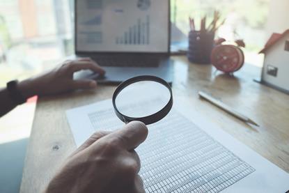 Scientist using magnifying glass to look through datasheet with laptop in the background