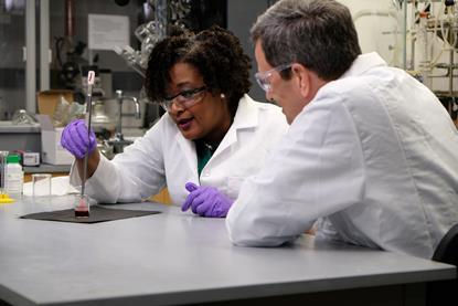 An image showing Malika Jeffries-EL with 'Beyond the Elements' presenter David Pogue. Both are wearing white lab coats. Malika is holding long tweezers with which she is pulling a stringy substance - nylon - out of a beaker filled with a reddish liquid.