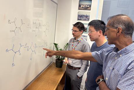 Three men discussing chemical structures drawn on a whiteboard