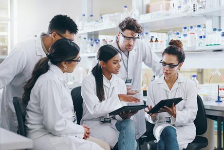 A group of young male and female scientific researchers in a lab and wearing lab coats and safety glasses gather around to discuss something on a computer tablet.