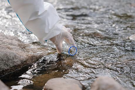 A photograph showing a gloved hand holding a vial to collect a water sample from a river