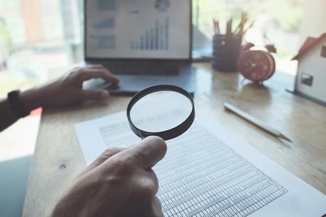 Scientist using magnifying glass to look through datasheet with laptop in the background