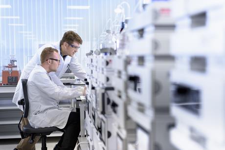 A photo of two white male scientists in labcoats sitting. One of them is sitting on a chair, the other standing next to him. Both are intently staring at a screen sitting on a lab bench.