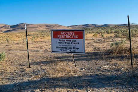 A landscape of dry mountainous grassland with clear blue sky. A sign in on a barb wire fence in the foreground says: Access Restricted/ Active Mine Site/Thacker Pass Project/ For access please call Lithium Nevada Corp