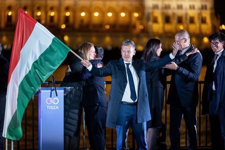 Peter Magyar standing on a stage waving a Hungarian flag while his supporters embrace each other behind him. On the other side of the River Danube you can see the Hungarian parliamentary building lit up at night.