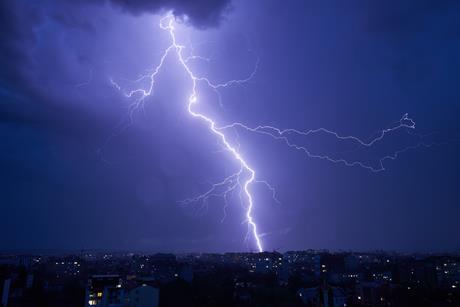 Powerful bolt of lightning cutting through dark, stormy sky over cityscape at night. Lightning's bright illuminate scene, contrasting sharply with dark clouds and dim lights of buildings below