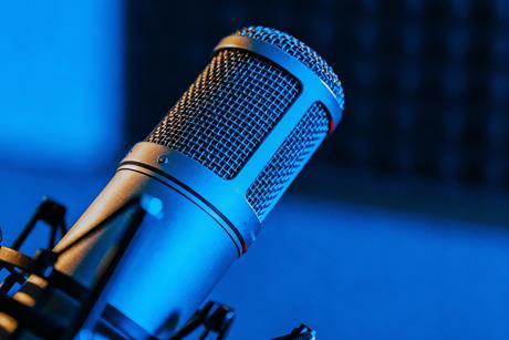 A detailed close-up of a silver microphone on a stand, positioned in a recording studio with a blue-lit background, showcasing its metal mesh grill and cylindrical body