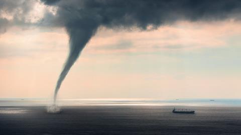 A photo of a waterspout, a tornado over water. There's dark clouds looming overhead and a large container ship appears tiny next to the enormous whirlwind extending from the clouds to the water surface.
