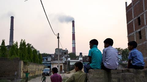 An image showing boys sitting and watching street cricket as emissions billow from smokestacks at the NTPC Ltd. Badarpur coal-fired power plant near residential property in Badarpur, Delhi, India
