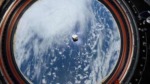 A photo of a tiny piece of gray rock floating in the centre of a circular window behind which is seen a space-view of Earth.