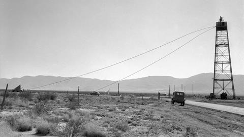 A black-and-white photo of the Trinity test site in 1945. It shows a desert landscape, scarce vegetation and a mountain range in the distance. On the right, there's a steel tower similar to an electric pylon but with a platform on top.