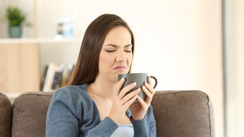 A white woman with brown hair wearing a white shirt and blue cardigan, sitting on a brown sofa chair. She's smelling a cup of coffee, a deeply disgusted look on her facecoffee