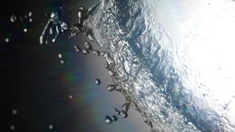 A backlit, closeup photo of a drop of water as it hits a surface, viewed from below. The drop appears as large circle covering most of the photo, with smaller droplets scattering away from it.