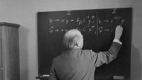 A black and white photo showing an older white man dressed in a suit standing in front of a blackboard with his back to the camera. His right arm is raised, he's holding a piece of chalk and is in the process of writing down equations on the board.