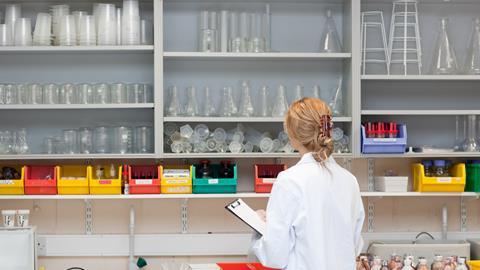 A photo showing a white blonde woman in a white lab coat. She's standing with her back to the camera in front of a lab bench, holding a clipboard on her left hand, inspecting at a wall full of shelves of glassware and other lab equipment