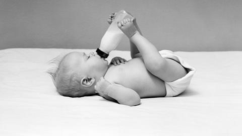 A black and white image showing a baby lying on his back and feeding himself from a bottle held by his legs