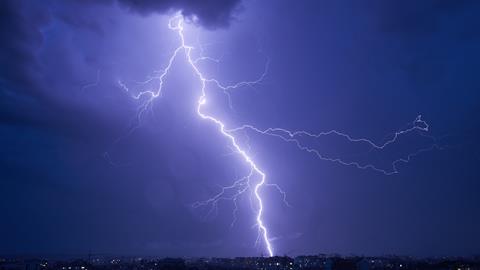 Powerful bolt of lightning cutting through dark, stormy sky over cityscape at night. Lightning's bright illuminate scene, contrasting sharply with dark clouds and dim lights of buildings below