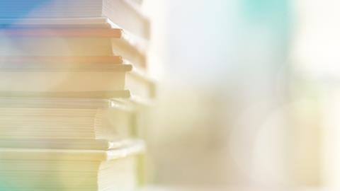 Shallow depth of field image showing close up of books piled high on desk and artificial lens flair obscuring dreamy style background