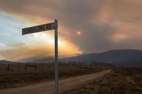 A road sign for Bumbalong Road by a track through the Australian outback in the distance heavy smoke from wildfires obscures the sun