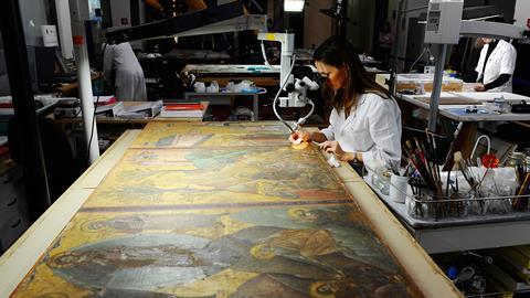 A photo showing a woman wearing a white lab coat looking through a miscroscope to inspect a section of what looks like a large medieval religious painting