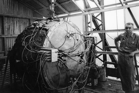 A black-and-white photo showing Norris Bradbury next to the Trinity test atom bomb. The device consists of a metal casing, a bit taller than Bradbury, with cables coming out of various places.