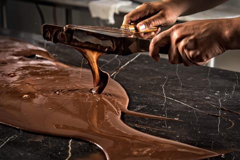 A photo of a person's hands holding two long spatulas covered in melted chocolate. The chocolate is dripping off the spatulas and pooling on a marble surface underneath, which is covered in more molten chocolate.