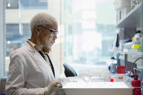 An image showing a Black woman with short white hair in a white lab coat, lab specs and purple plastic gloves sits at a bench focusing on a number of sample tubes in front of her