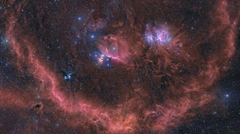An image showing the Orion stellar neighbourhood as red-glowing dust clouds and hundreds of tiny dots of newly born stars