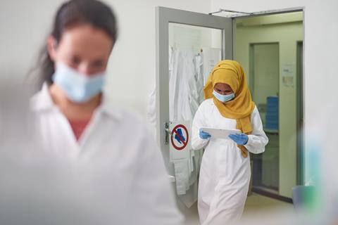 A woman wearing a headscarf and lab coat entering a laboratory through a door while reading a tablet