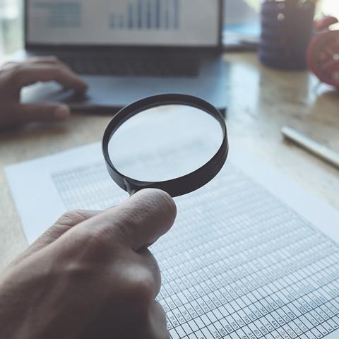 Scientist using magnifying glass to look through datasheet with laptop in the background