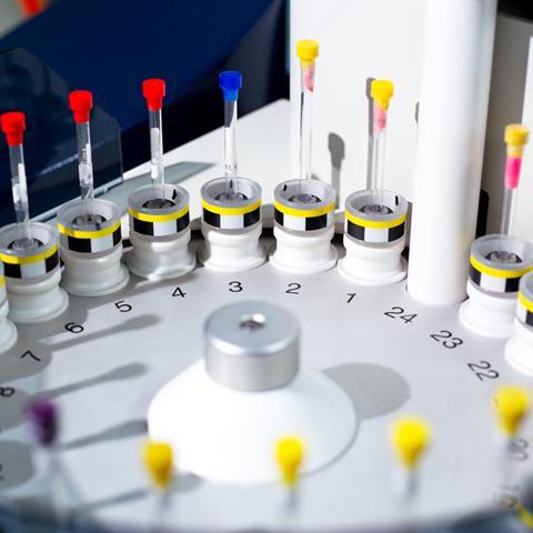 Close-up of a laboratory sample analysis machine with multiple vials in a circular arrangement, featuring color-coded caps in red, yellow, and blue. The equipment is used for automated testing or sample processing in scientific or medical research