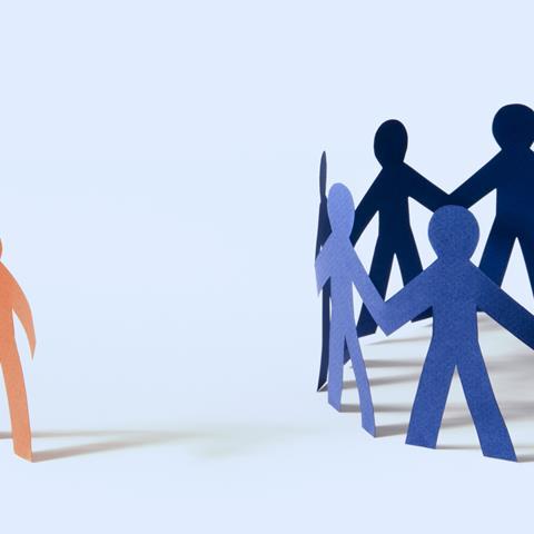 A photo of a of paper cutouts in the shape of a person. A number of blue ones are stood in a circle, next to them, slightly apart, is a single red one.