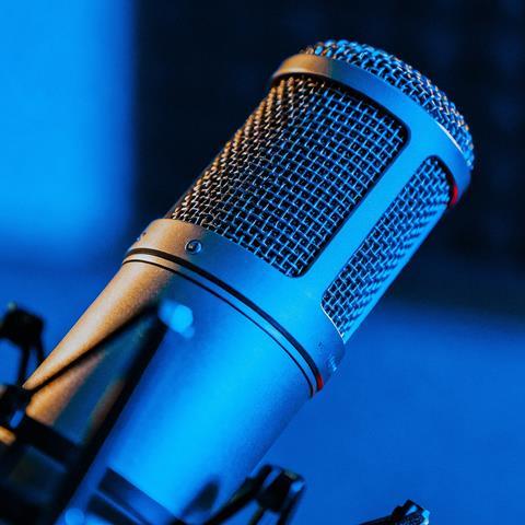 A detailed close-up of a silver microphone on a stand, positioned in a recording studio with a blue-lit background, showcasing its metal mesh grill and cylindrical body