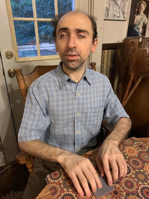 A photo of a white man with dark hair and stubbly beard wearing a short-sleeved checkered shirt. He's sitting at a table, his fingers resting on a palm-sized lithophane image of a protein electrophoresis gel.