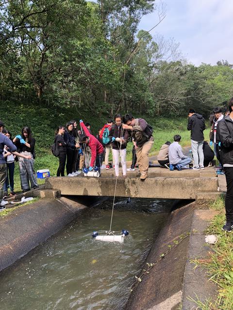 A group of young adults stand around a waterway. Two people on a concrete platform over the water hold a string connected to a plastic device in the water that is analysing the water