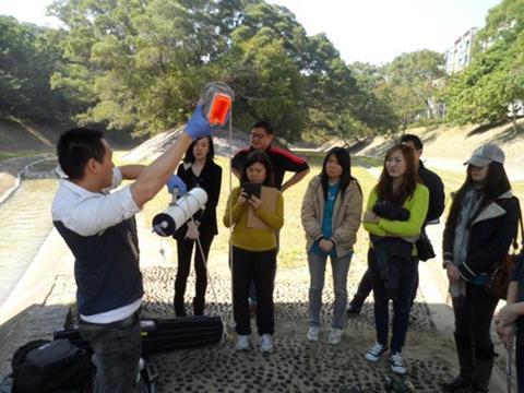 A man holds up a device for monitoring water quality watched by a group of 7 people