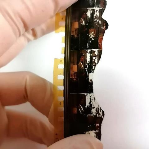 Close up of a damaged strip of celluloid film held by a hand in latex gloves. The film features two women in mid-century living room and clothing but the film is cracked and broken and parts of the images have turned white.