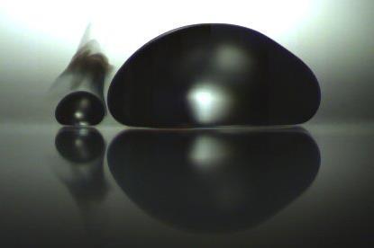A photo of two droplets sitting on a surface, one larger and one smaller. They are backlit so they appear as shaded outlines. Between the droplets, a wisp of evaporating liquid can be seen