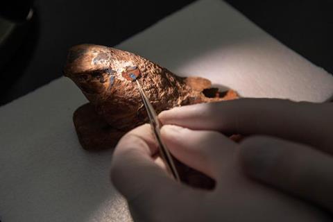 Hands in latex gloves use tweazers to remove a blue pigment from an old falcon figurine