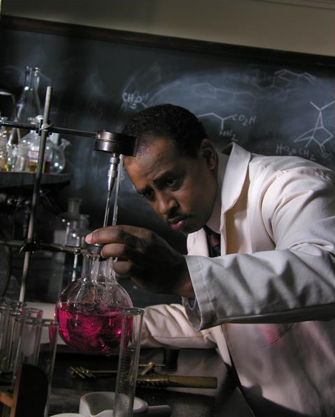 Actor Ruben Santiago-Hudson as Percy Julian in the documentary Forgotten Genius. He's in the lab, wearing a lab coat, bending down to examine a round bottom flask containing a pink liquid.