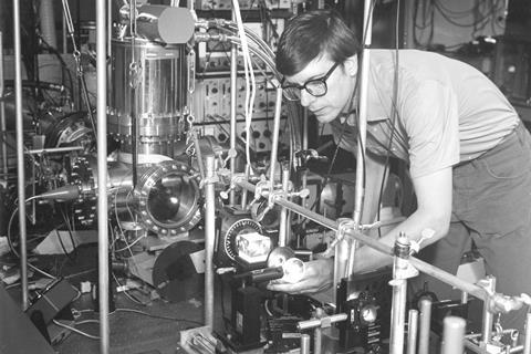 A black-and-white photo of a young man with thick-rimmed glasses bending over an array of tubes, steel rods and lenses
