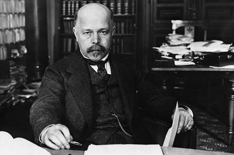 A black and white photo of well-dressed bald white man with a mustache and goatee in a messy office