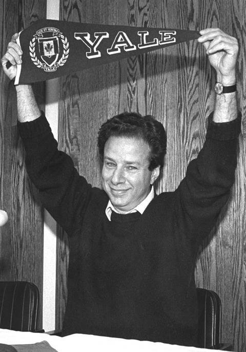 A black-and-white photo of a white man with dark hair dressed in a dark jumper. He looks excited and happy, holding a smaller banner reading 'Yale' above his head.