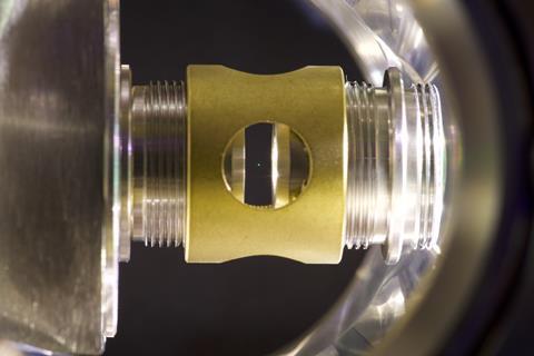 A photo showing a metallic golden apparatus with a gap in the centre. In this central gap there is a tiny green dot - the glass sphere