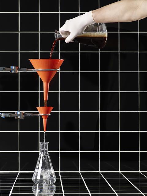 A picture showing a man wearing a glove and pouring liquid through funnels into glass flask